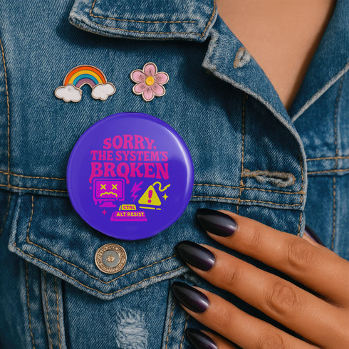 A vibrant purple pinback button with a glossy finish. The main text reads "SORRY, THE SYSTEM'S BROKEN" in a chunky, dripping pink font. Below the text are several retro tech icons in pink and yellow. On the left is a glitching 90s-style computer monitor with a sad face and X's for eyes. In the center is a yellow warning sign with an exclamation point, a lightning bolt, and sparkles. Below is a keyboard command parody that reads "CTRL ALT RESIST.